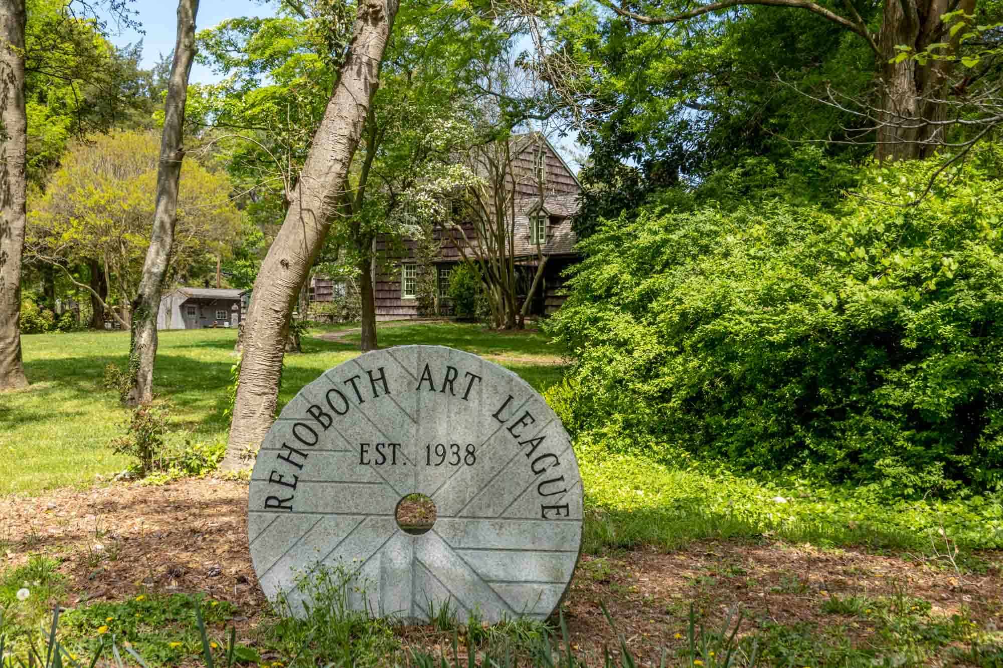 Circular sign: "Rehoboth Art League, Est. 1938) amidst trees and plants