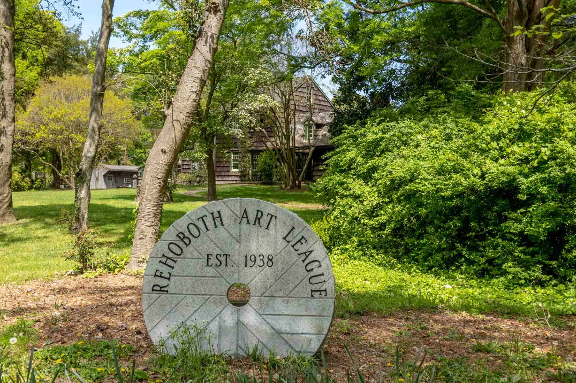 Circular sign: "Rehoboth Art League, Est. 1938) amidst trees and plants.