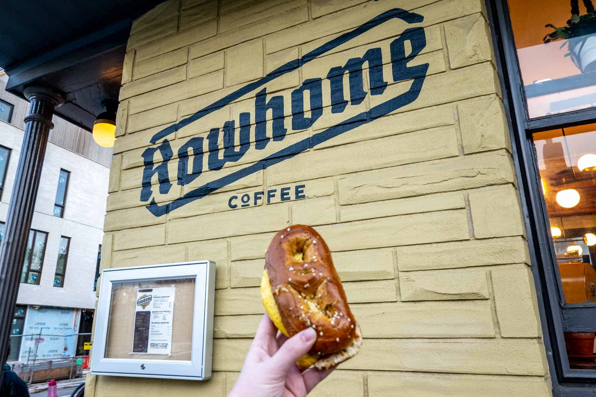Hand holding a pretzel sandwich in front of a sign for "Rowhome Coffee"