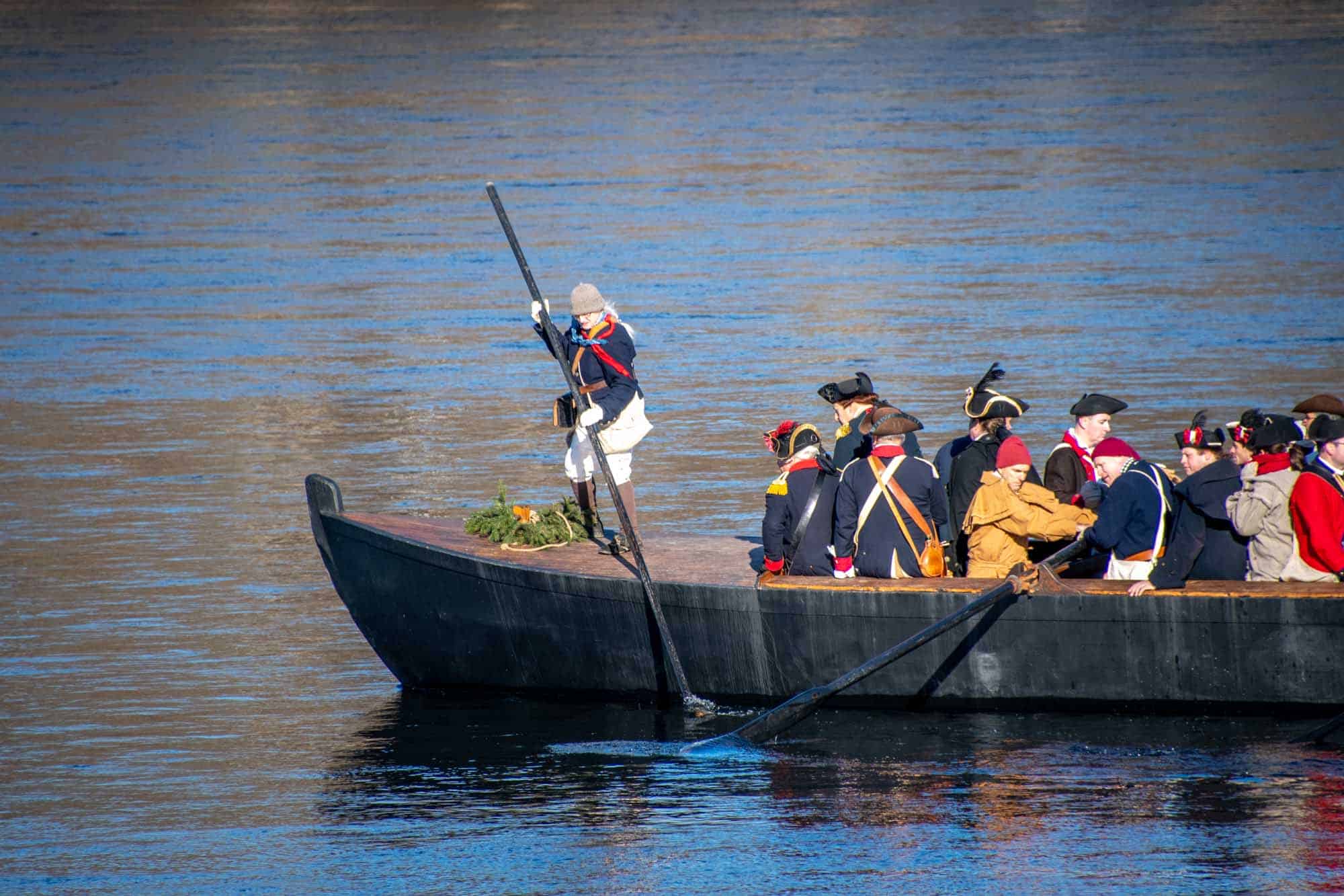 Person in Colonial dress standing on the bow of a wooden boat next to other "troops" in the boat