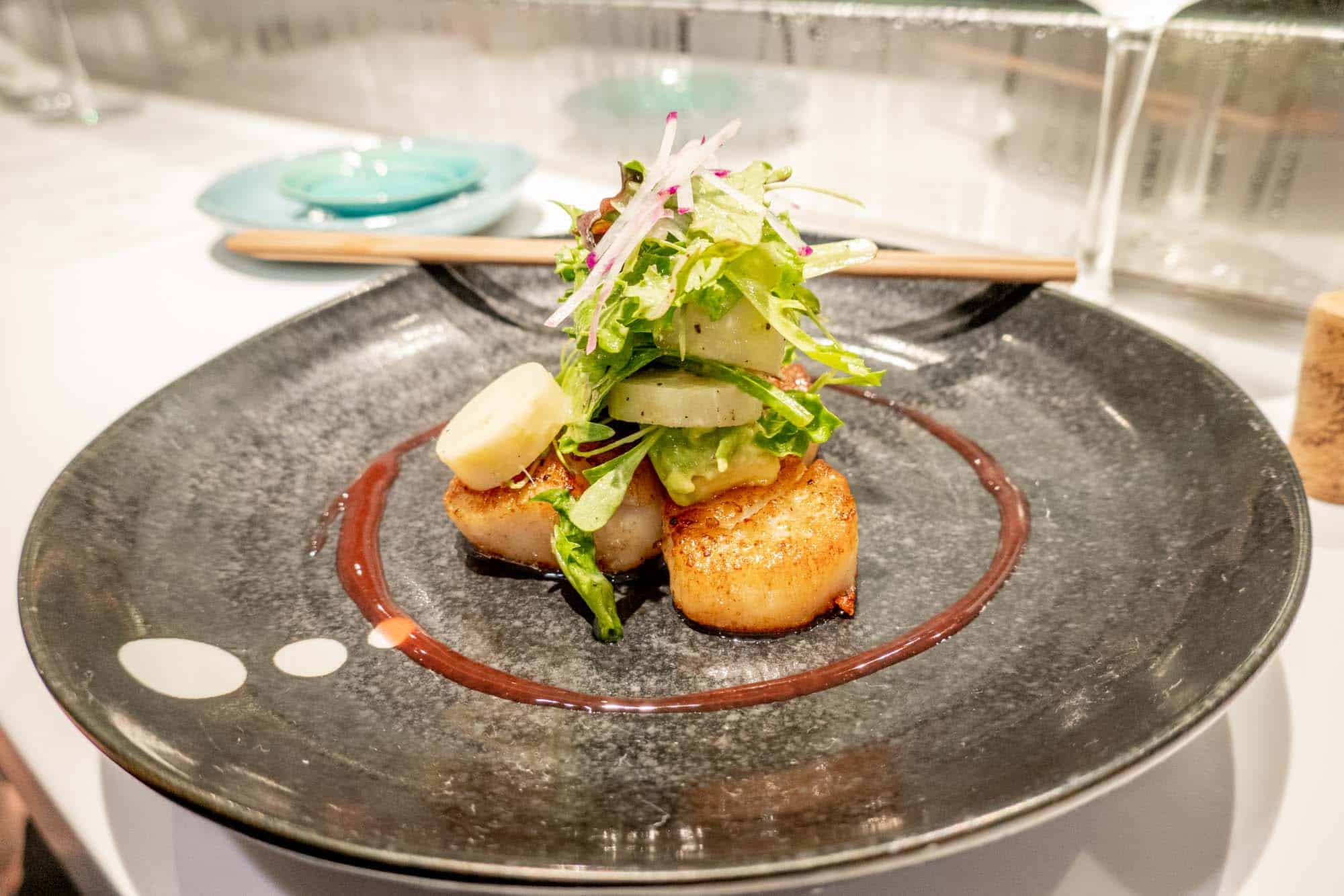 Scallops topped with a salad on a black plate with chopsticks