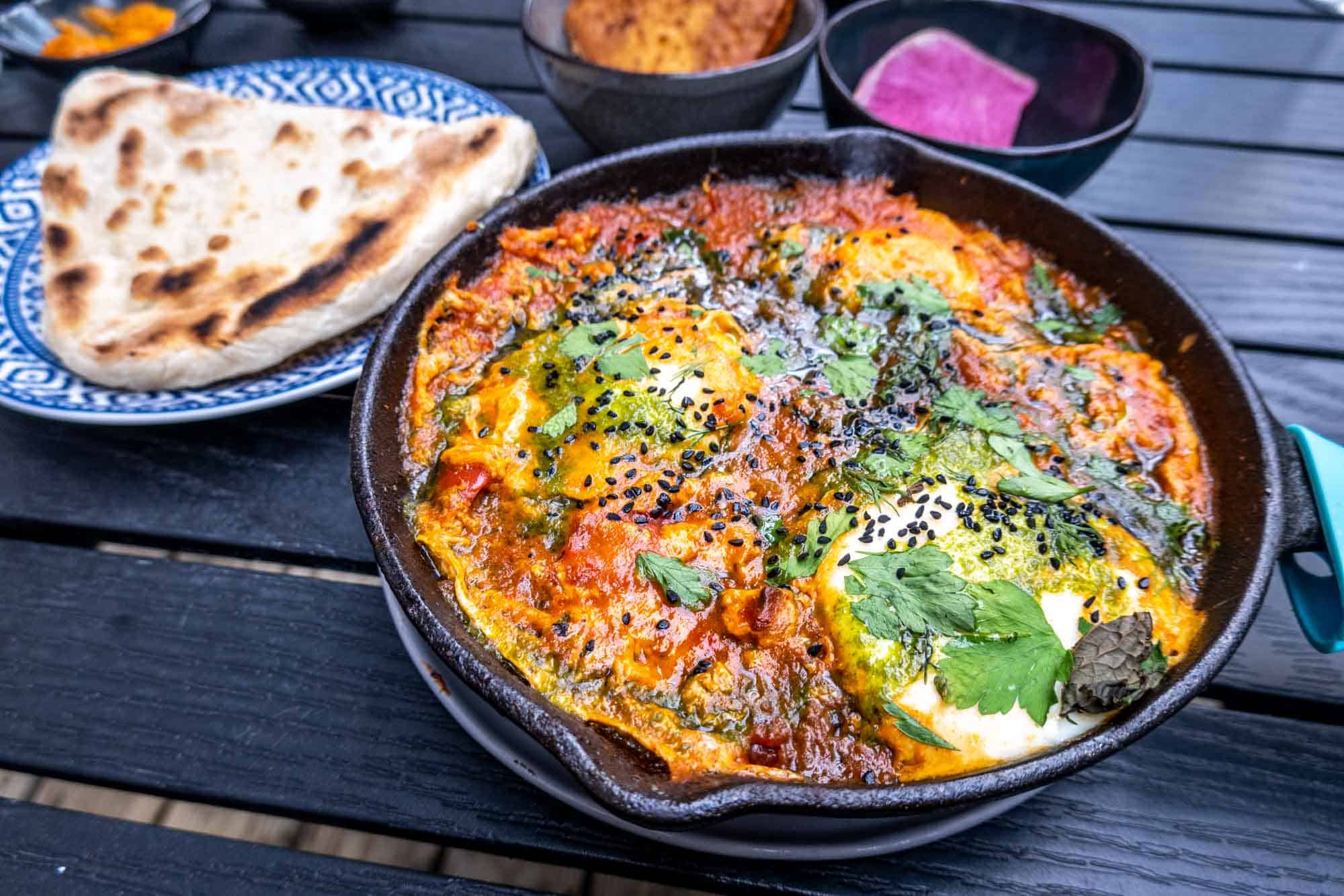 Cast iron skillet filled with shakshuka and eggs beside a flatbread.