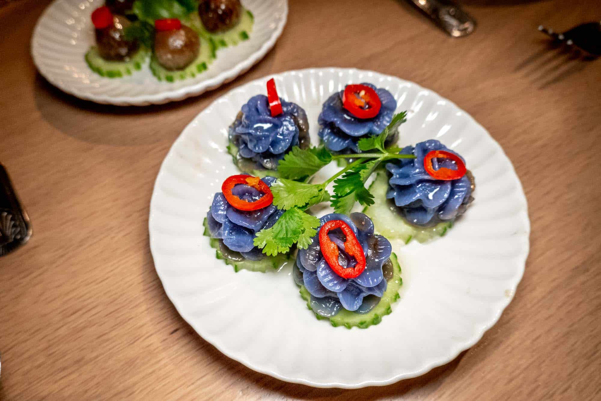Purple flower dumplings on cucumber topped with red pepper on a white plate.