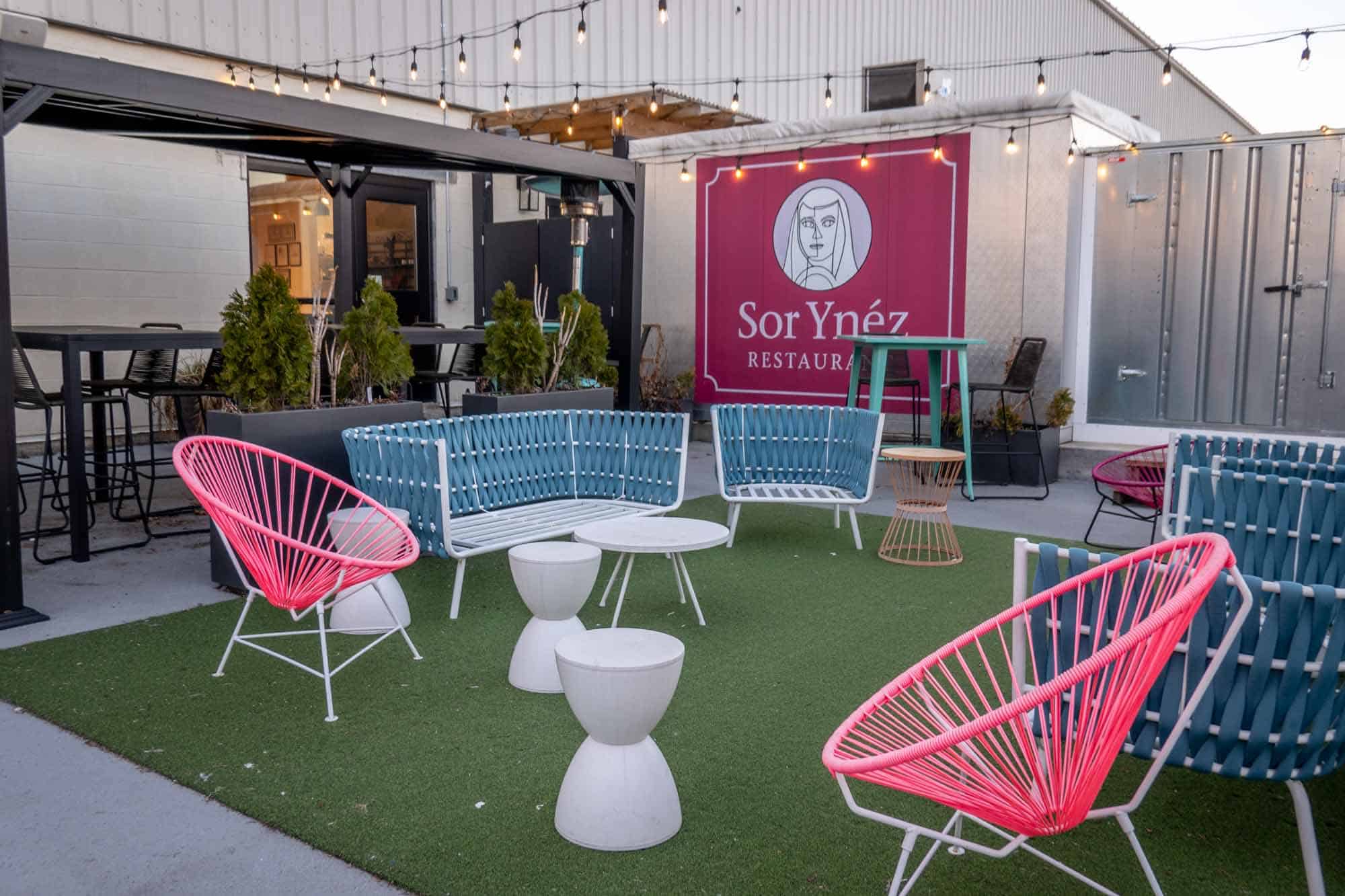 Bright blue and pink chairs on green AstroTurf in front of 'Sor Ynez Restaurant' sign