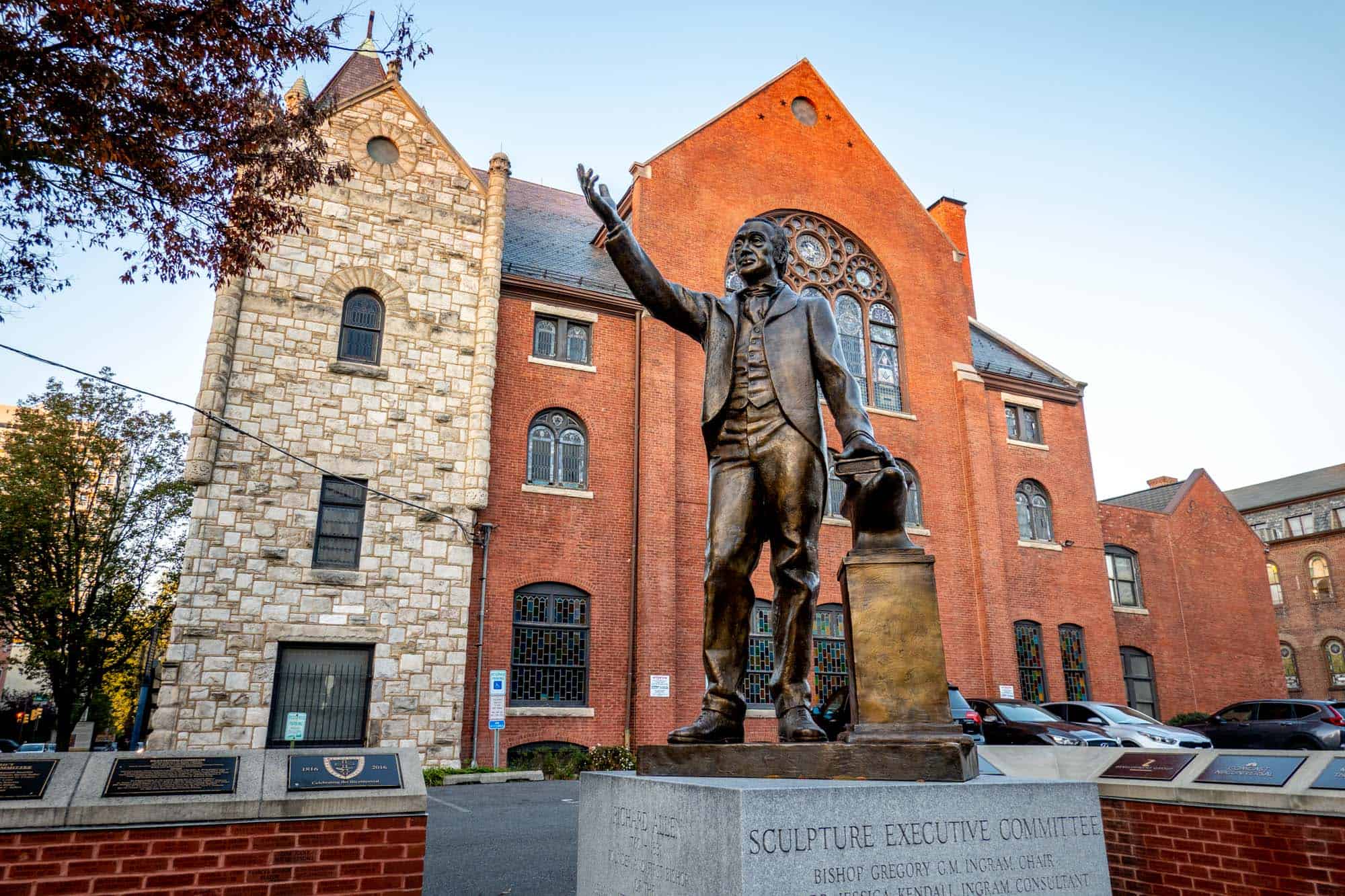 Bronze statue of a man with his arm raised outside a red brick building with stained glass