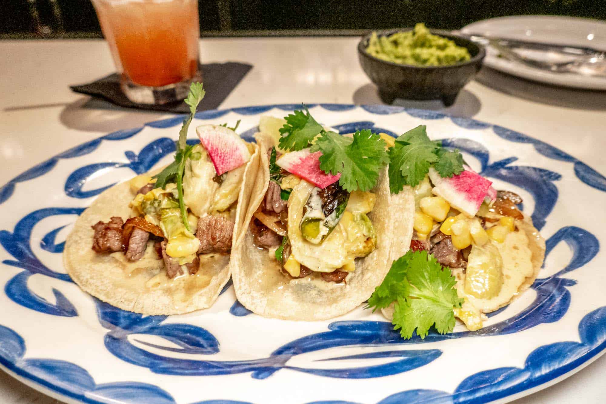 Three steak tacos on a plate with a drink and guacamole in the background