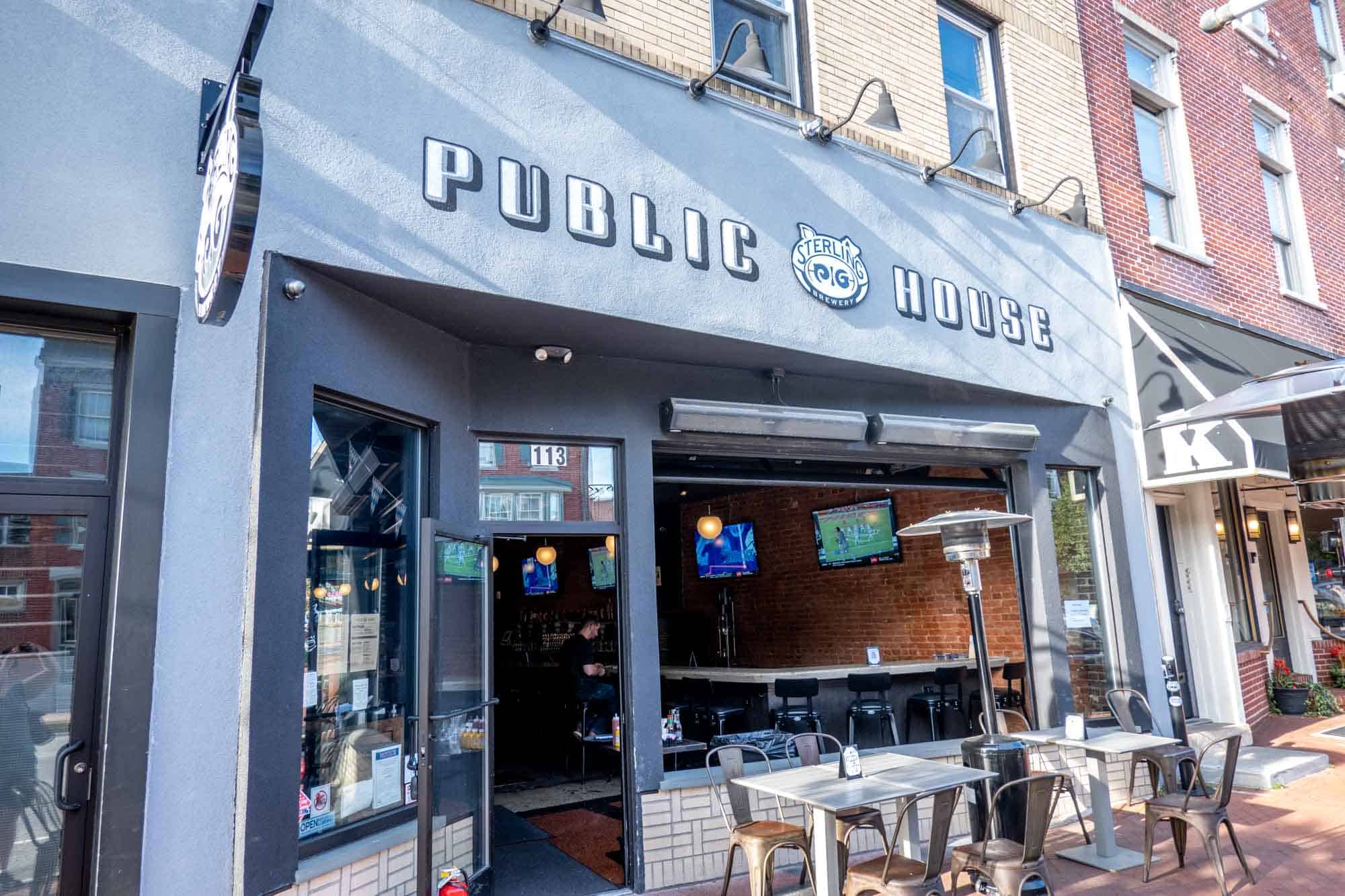 Table and chairs on the sidewalk in front of a gray storefront and a sign for "Sterling Pig Public House."