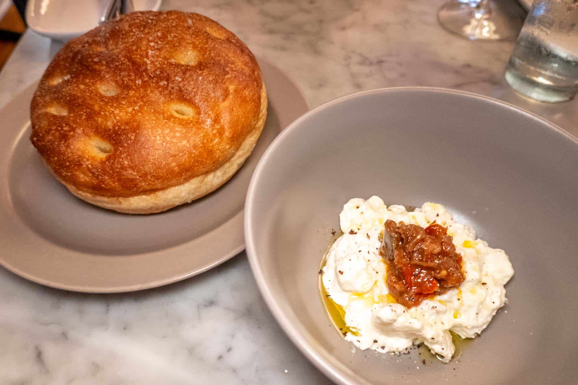 Bowl of stracciatella cheese along with a plate containing a large loaf of focaccia bread