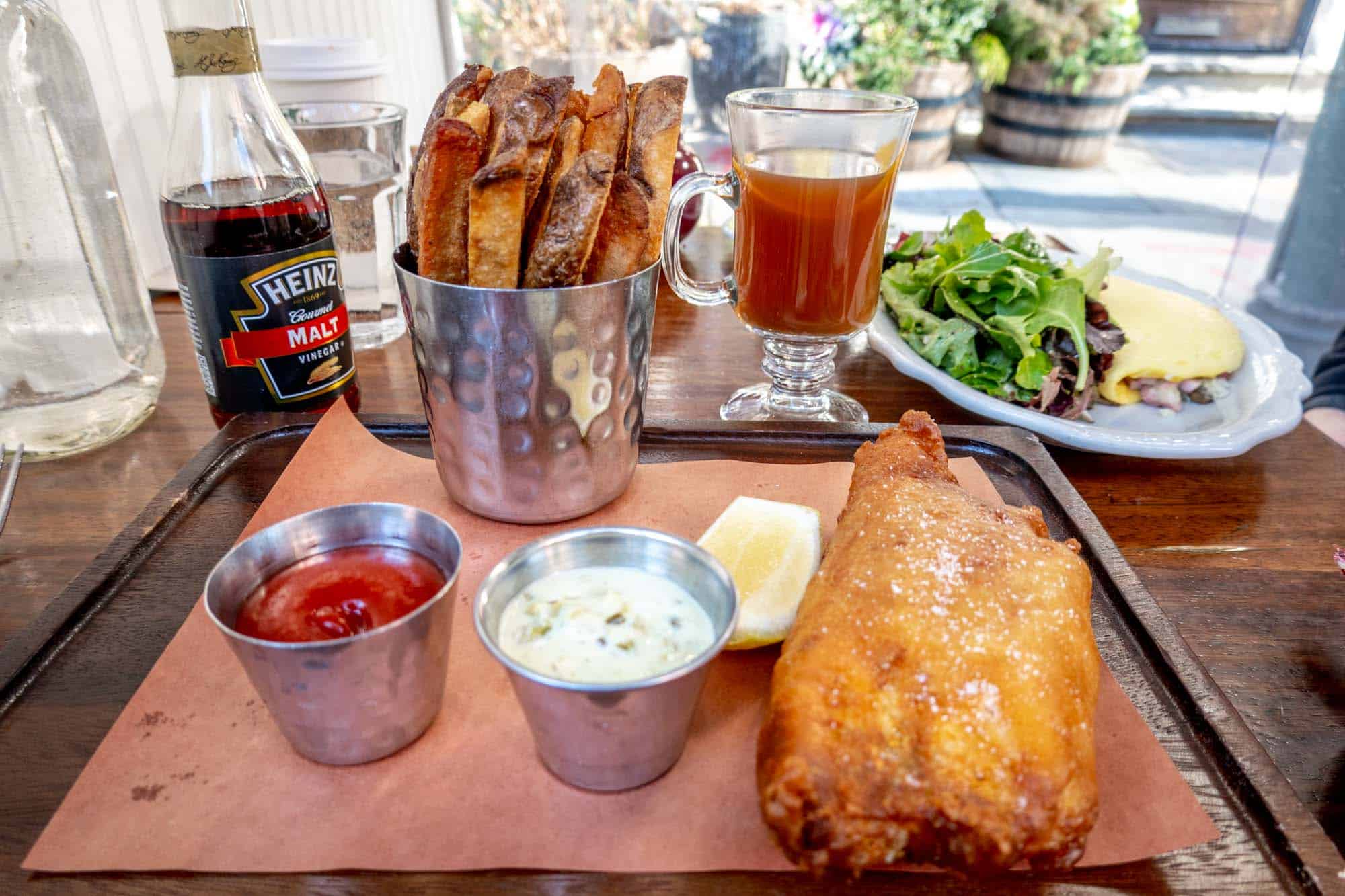 Fish and chips on a wooden platter with a hot toddy cocktail and omelet in the background