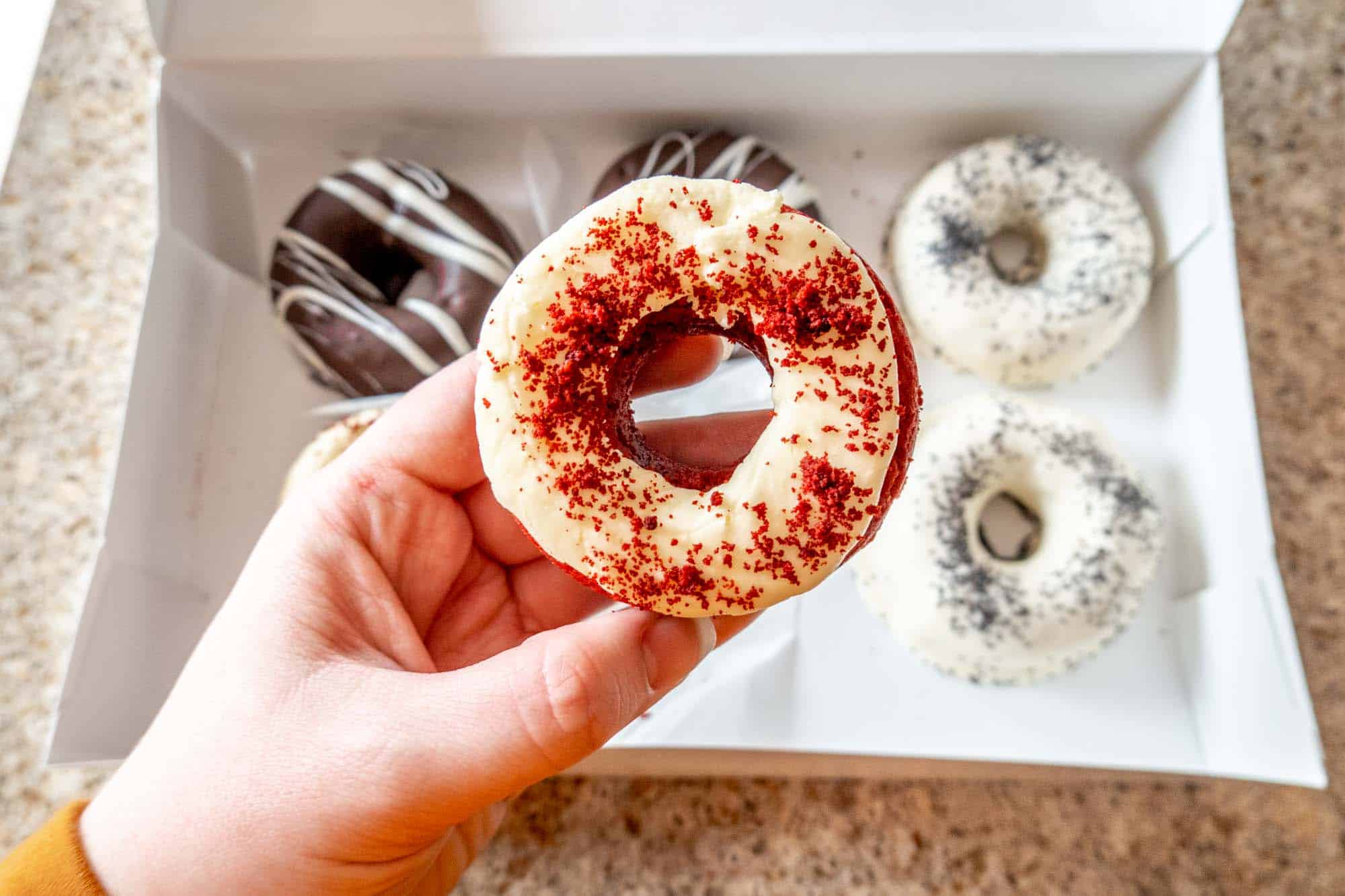 Hand holding a red velvet donut with a box of a half-dozen donuts in the background.