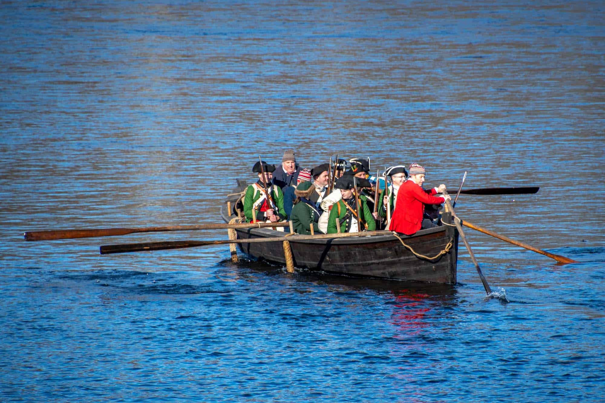 Reenactors in Revolutionary era dress rowing a wooden boat across the Delaware River