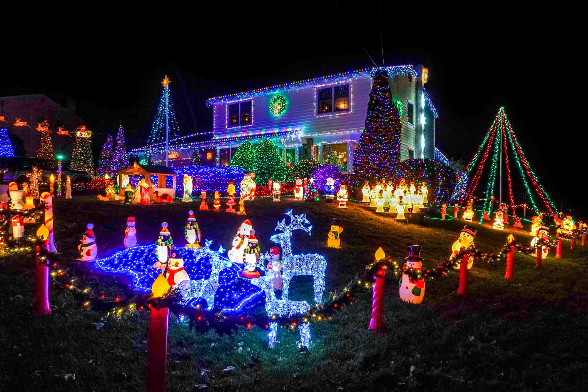 Yard packed with Christmas and winter illuminated figurines beside a house covered in lights.