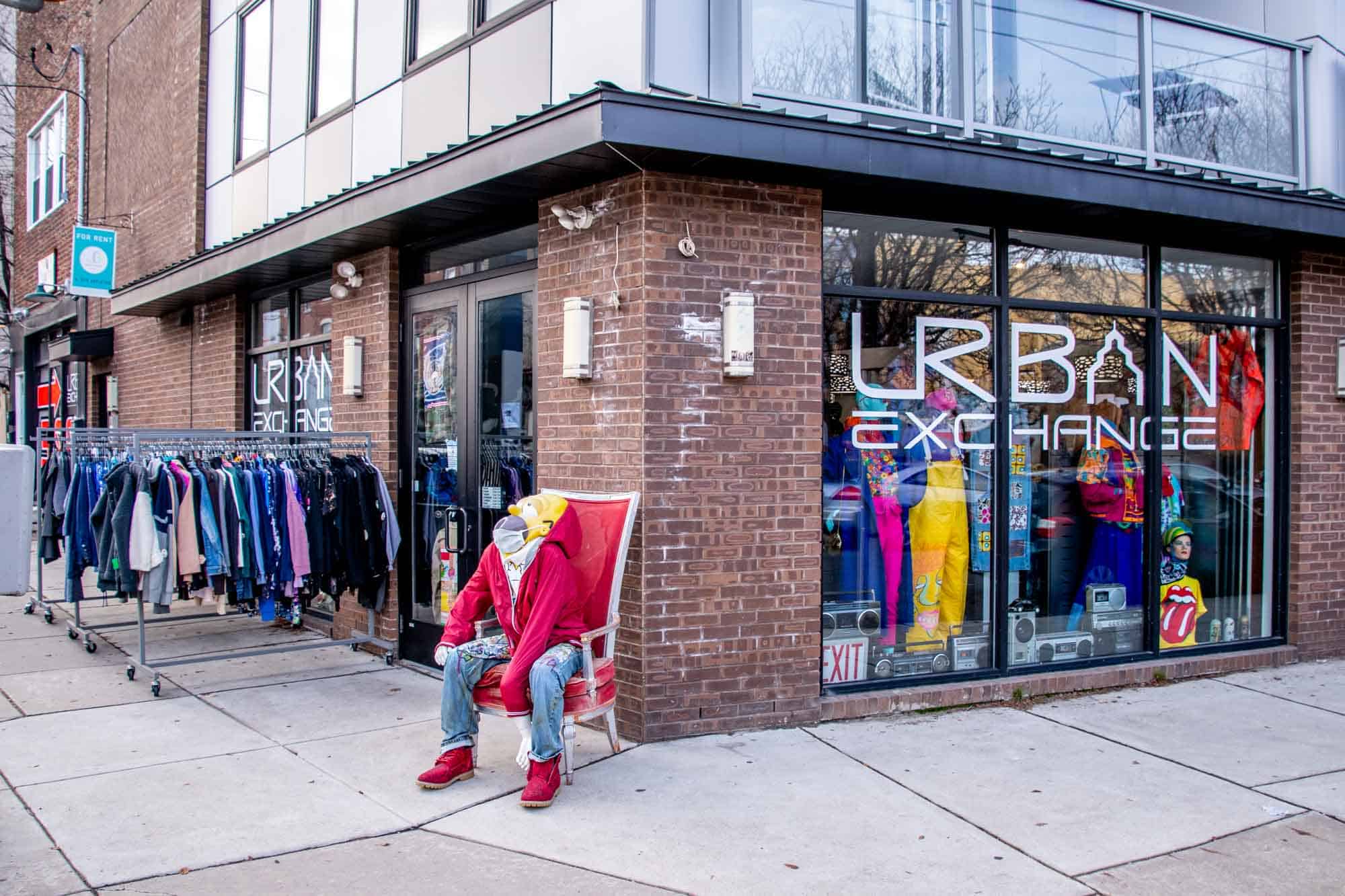 Racks of clothes and a mannequin sitting in a chair outside the brick exterior of a store called Urban Exchange.