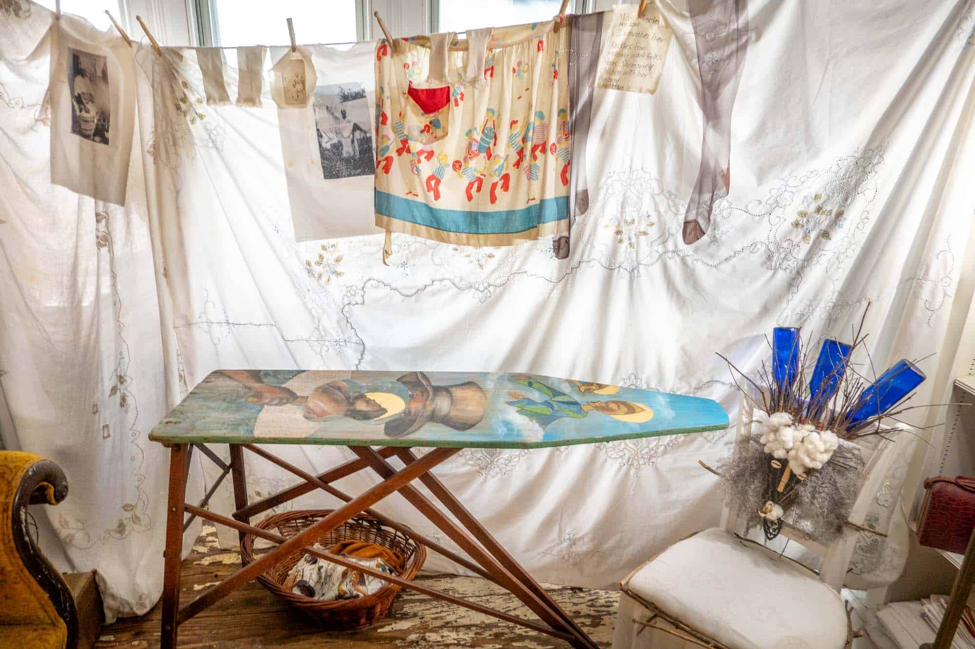 Clothes and a tablecloth hanging from an indoor clothesline beside an ironing board and artwork.