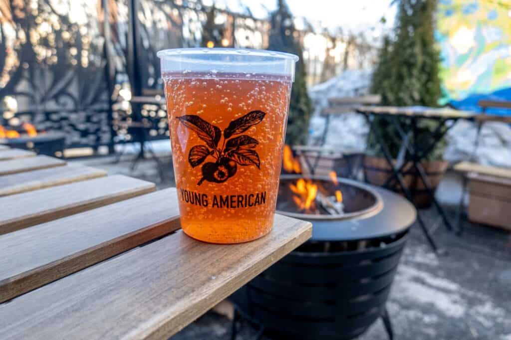 Glass of cider with a logo for "Young American" sitting on a table outside with a fire pit in the background.